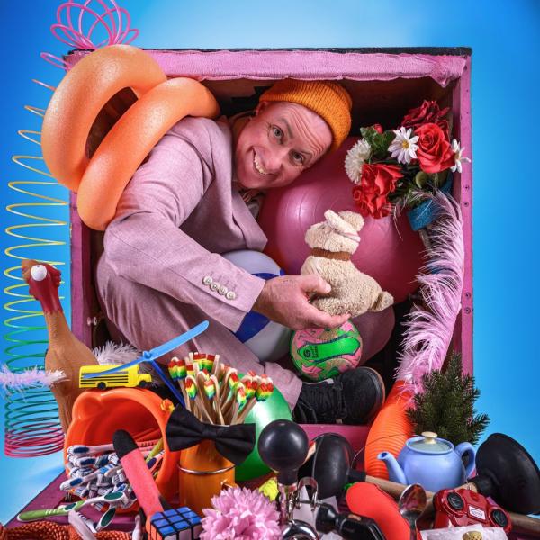Man cramped inside of a pink box alongside various toys and other objects that are spilling out of the box like an exercise ball, slinky and squeaky chicken. He is holding a toy stuffed dog.