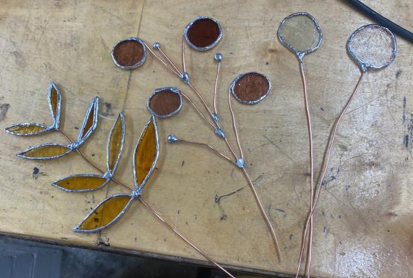 A photograph of three flower stems made of silver wire and stained glass. These are laid upon a wooden workbench