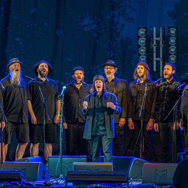 The Spooky Men's Chorale sing into microphones together on stage, in dark outfits and hats, lit by a blue stage-light.