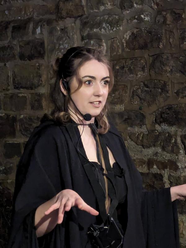 A white, female tour guide with brown hair, a black cloak and a microphone gestures with a ghoulish expression as she tells a scary story. She is stood in front of a dark stone wall at night time.