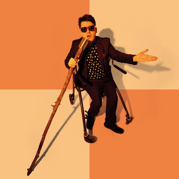 Jack Carroll in a black suit, polka dot shirt and sunglasses speaks into a tin can on a long wooden stick, his other hand outstretched, positioned in front of a orange and yellow chequered pattern.