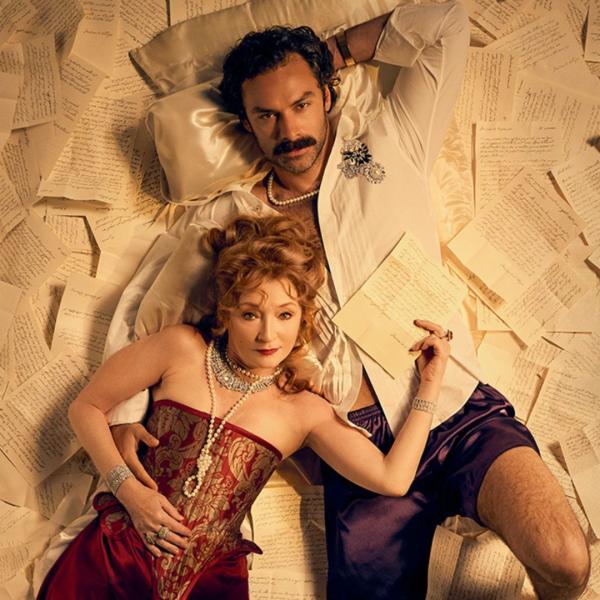 A woman in pearls, diamond jewellery and a red and gold corset lies on a moustached man in an unbuttoned shirt and shorts. Both look up at the camera and lie across sheets of handwritten notes. 