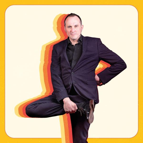 Phil Ellis stands upright, one hand on hip and his leg raised to his hip. He wears a dark suit and is surrounded by a silhouette of yellow, orange and red.