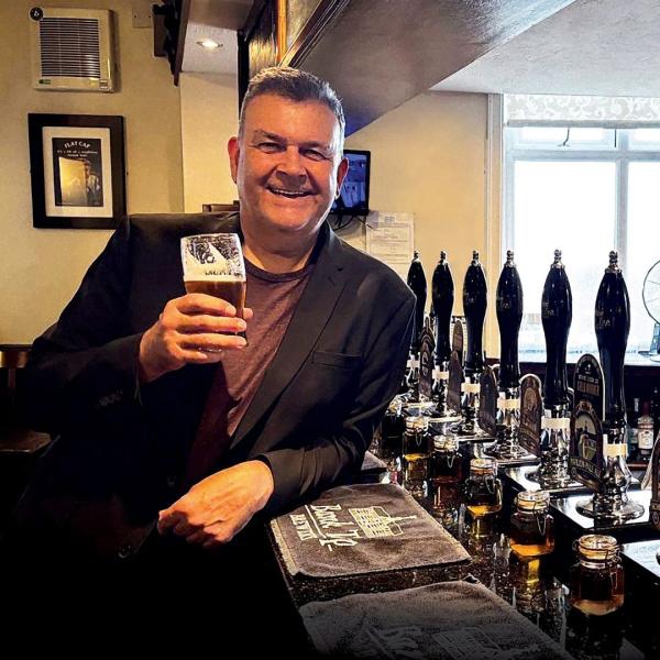 A man stands beside a bar in a pub, cheerily holding up a pint of beer. 