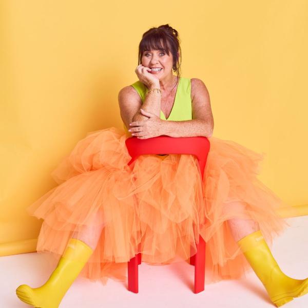 Coleen Nolan sits playfully on a red plastic chair, in yellow wellies and a pale orange tutu, all set against a yellow backdrop. Her arms are perched on the chair and she cheekily rests her face in her palm.