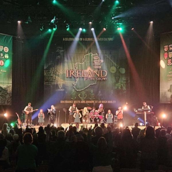 A large band with multiple instrumentalists and singers on stage perform under spotlights to a large crowd.