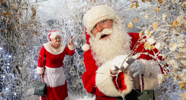 A snow filled scene with Santa and Mrs Claus. Santa is dressed in his red coat with white fur cuffs and red hat with white fur cuffs with a long white beard and white gloves stands smiling. Mrs Claus is in the background looking forward, holding a Christmas Experience branded gift bag. She is dressed in a long red dress and bonnet both with white fur trim.
