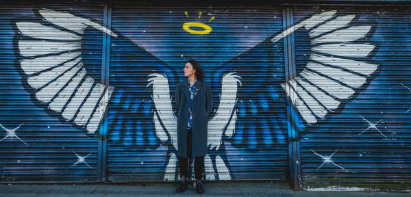 A man in a long grey coat stands in front of a blue shutter door painted with large white wings and a yellow halo, making it look like he has wings.