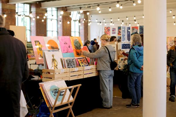 A visitor inside the busy Print Fair at last year's festival inside the 1912 Mill. The space is a former textile mill with red brick walls, white pillars and strings of festoon lights hanging from the high beams. The space is filled with stalls of original prints and handmade books.
