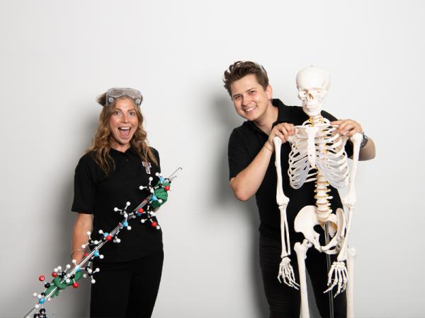 Two people holding scientific objects, one skeleton and one dna stick smiling at the camera.