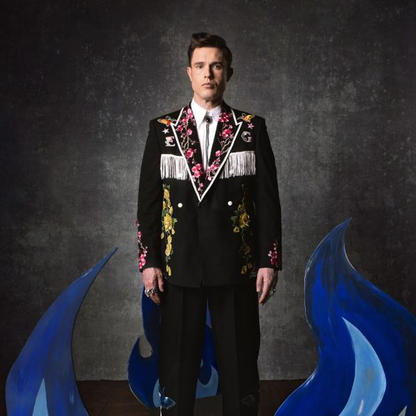Ed Gamble stands up straight with a neutral expression, wearing an elaborate patterned suit jacket with flowers and tassels, in front of a textured grey backdrop and blue flame.