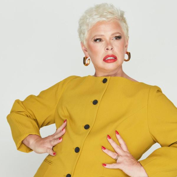 Laura Smyth looks fiercely off-camera, hands on her waist and is wearing a yellow button-up dress, red lipstick and red nails.