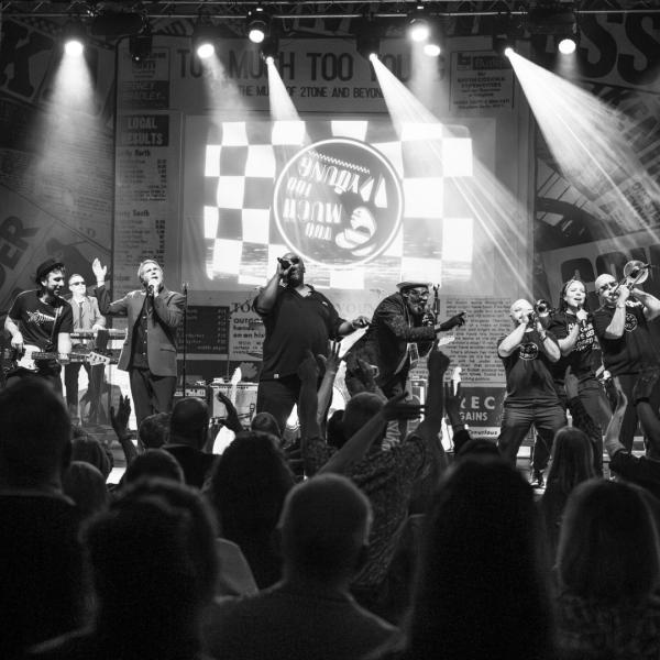 Black and white shot of a band performing on stage, including three vocalists at the centre and several band members playing trombones and trumpets.