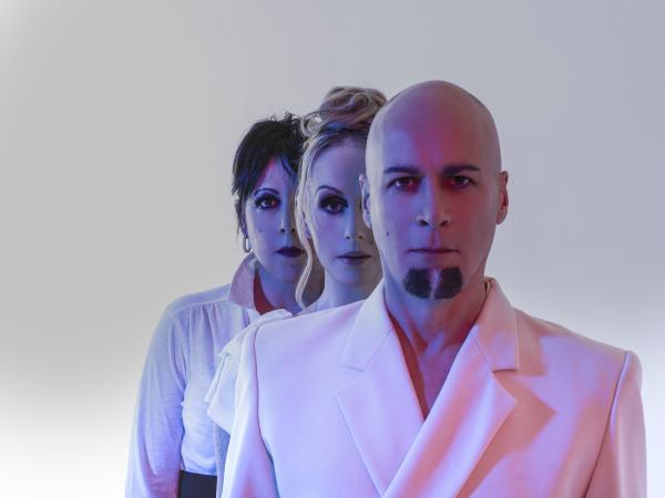 Three people in white clothing stand in a line, facing forward with serious expressions against a light background with blue and pink lighting.