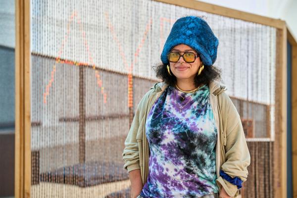 A person stands smiling in front of a large woven installation framed in wood. They wear a bright blue fuzzy hat, oversized tinted glasses, large gold hoop earrings, a nose ring, layered necklaces, a colorful purple, blue, and black tie-dye shirt, and a light beige jacket. Their hands are tucked into their pockets. Behind them, the woven artwork partially reveals glowing orange letters, creating a textured backdrop in a bright gallery space.