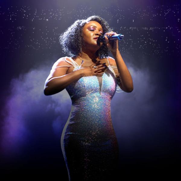 A tribute act of Whitney Houston wearing an iridescent dress, singing into a microphone against the backdrop of a dark auditorium.