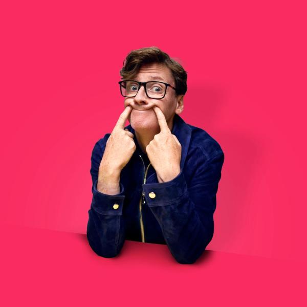 Ed Byrne poses, poking his fingers into his face to form a humorous forced smile. The photo is against a pink background while he leans on a pink table that blends in.