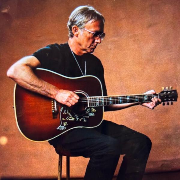 Steve Cradock sits on a stool and plays a patterned acoustic guitar, wearing a black t-shirt and sunglasses.