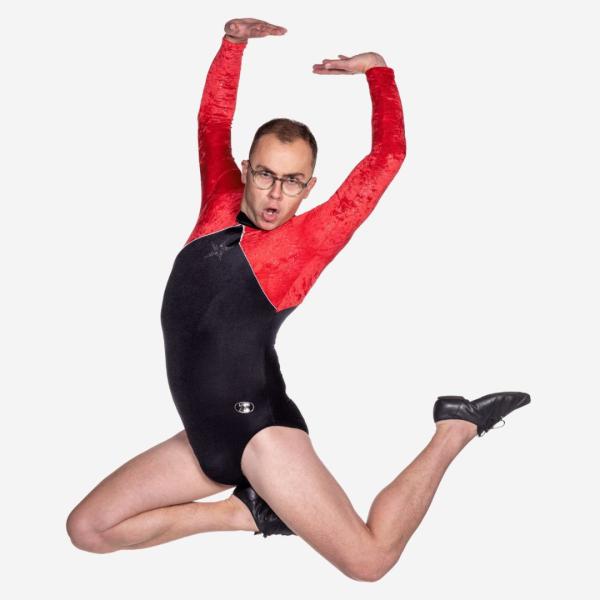 In a black and red leotard, Joe Tracini strikes a pose in mid-air with arms and legs outstretched, with an open-mouthed expression.