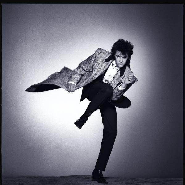 Old photo of Paul Young standing on his left leg with his left knee up by his chest. He has a dark 70s-style haircut, wearing a long coat, white shirt and black trousers.