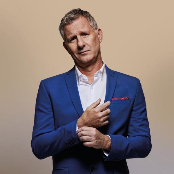 Adam Hills stands wearing a blue suit and clown shoes.