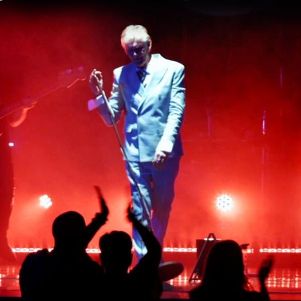 On a hazy stage with red spotlights, a David Bowie tribute act performs in a white suit, accompanied by two guitarists. 