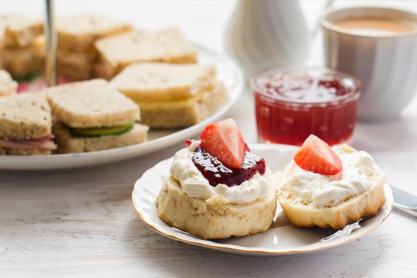 Scones with cream, jam and strawberries, with a plate of sandwiches behind