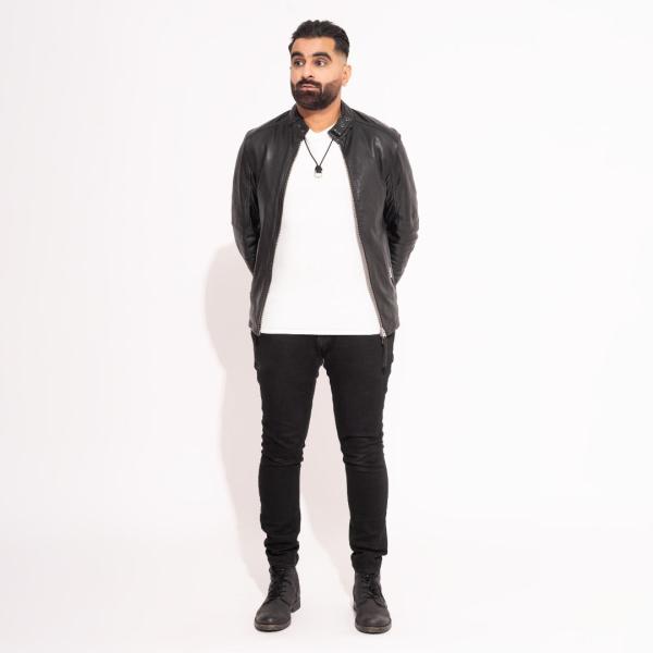 Tez Ilyas stands with his hands behind his back, wearing a leather jacket and white t-shirt as he looks off-camera in front of a white backdrop.