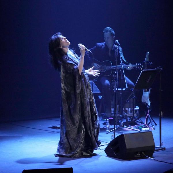 In moody blue lighting, Imelda May passionately sings on stage in a flowy dress, in front of an acoustic guitarist