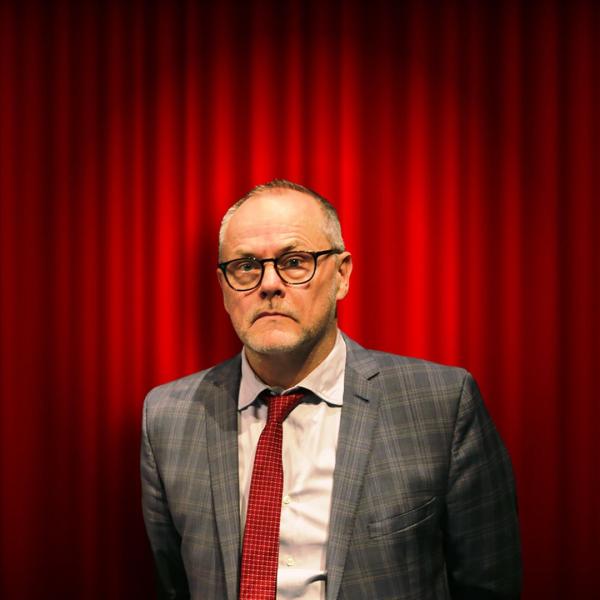 Jack Dee holds a neutral expression, wearing glasses, a grey suit and red tie, positioned in front of a red curtain and lit by a spotlight.