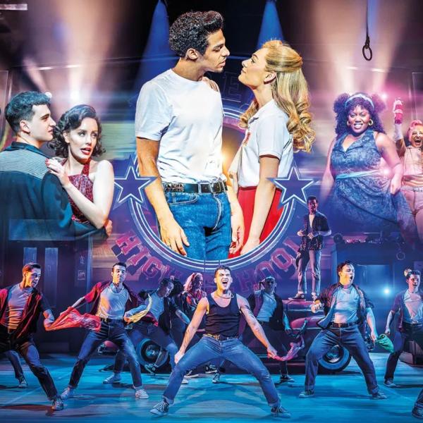 A young man with a greased quiff and white t-shirt tucked into denim jeans looks into the eyes of a blonde woman with a white collared shirt and red skirt. The couple are surrounded by the cast of Grease, jumping and dancing together in front of a vintage car and high school gym backdrop.