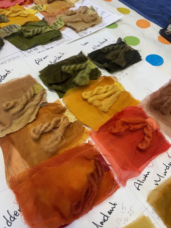 Squares of fabric and skeins of wool dyed in natural shades of ochre, orange, red and green, laid out on labelled paper
