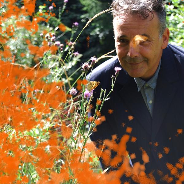 Rob Newman wearing a suit, obscured by some plants with orange flowers.
