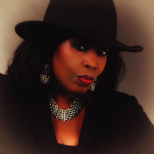 Ruby Turner posing in front of a plain backdrop, wearing silver jewellery, a black outfit and brimmed hat.