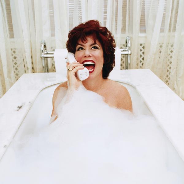 Ruby Wax smiles widely and speaks into a wireless telephone in a bubbly white bath tub, wearing striking red lipstick and make-up.