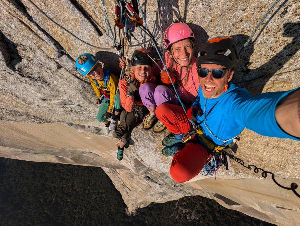 A family sit harnessed and perched on the side of a large cliff face, all are smiling up at the camera