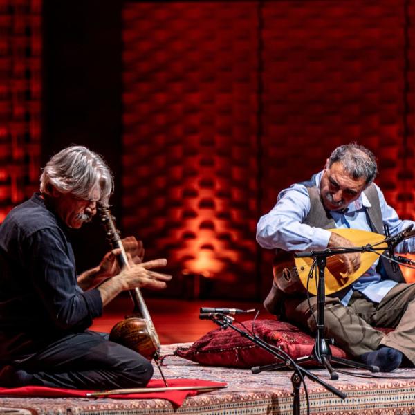 Kayhan Kalhor and Erdal Erzincan sit on cushions on a Persian rug, with their heads down as they play their instruments. They are in front of a red, textured background, lit by warm ambient lighting.
