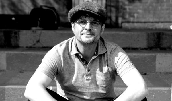 A man sits on some steps he is unshaven with round glasses and a baggy cap, similar to the baggy green of the Aussie cricket team, but like I say it's black and white so he probably not and Aussie cricketer what with him being a singer and all