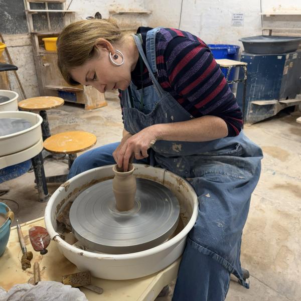 A woman is sat at a pottery wheel and is throwing a small vase
