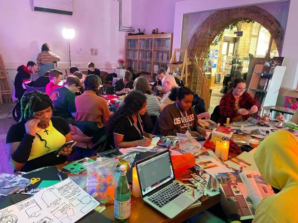 A busy room lit in pink. People are sat at tables covered with magazines and coloured paper as they create zines.