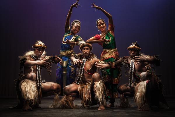 On a dark background, 5 dancers are posed in formation. They are wearing traditional South Asian and South African dress. The 3 males are crouched down on the ground with their arms outstretched in front of them. The 2 women are behind them, stood in the gaps of the men. They are leaning in towards the middle, their inner arms bent at the elbow and hands at chest-height. Their outer arms are lifted above their heads and reaching inwards.