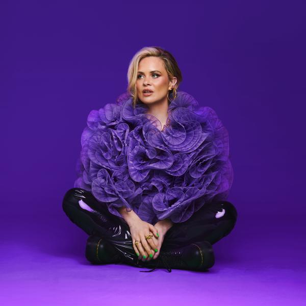 Joanne McNally looks to the side and sits cross-legged in shiny black trousers and an oversized, frilly purple top, in front of a purple background.