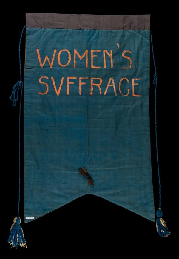 A hand made banner reads "Women's Suffrage" in orange writing on green fabric