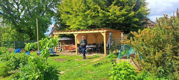 photograph of seacroft forest garden showing a wooden shelter in the background, a man standing in front of the shelter, surrounded by plants and trees.
