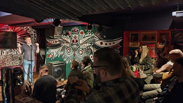 A male comedian on stage talking into a microphone to a crowd. The room is painted in bright Mariachi colours.