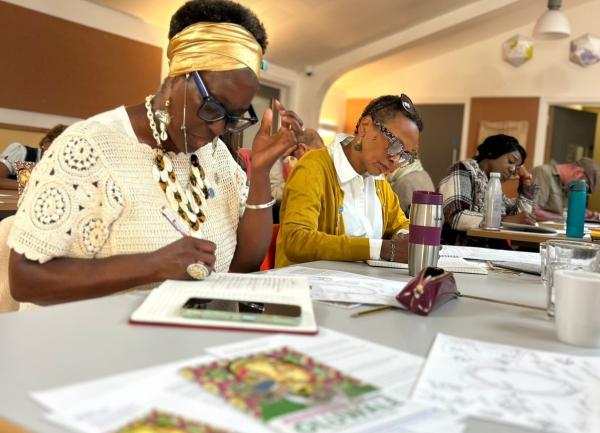 Participants at a creative writing workshop sit around tables, concentrating on their writing. In the foreground, a woman wearing a gold headwrap, glasses and a white crocheted top writes in a notebook, while others work on their writing in the background. Papers, notebooks and drinks are spread across the tables.