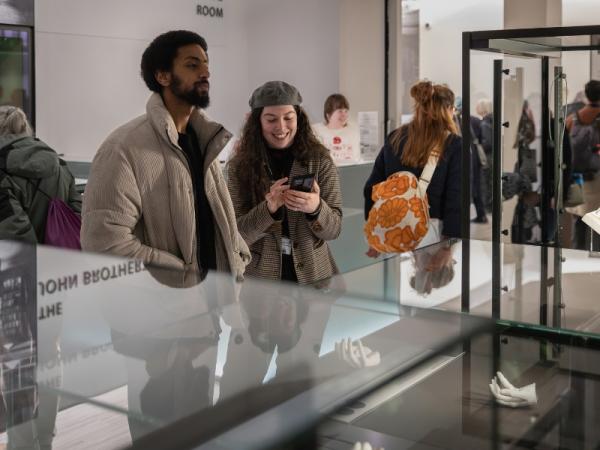 People stood in a museum surrounded by objects in glass cases. In the centre are two people stood together whilst one smiles at their phone.