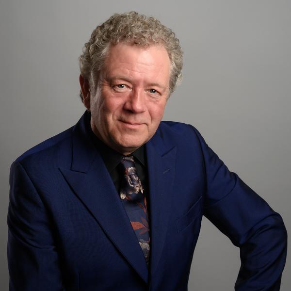 Jon Culshaw smiles in a navy blue suit and dark patterned tie. 
