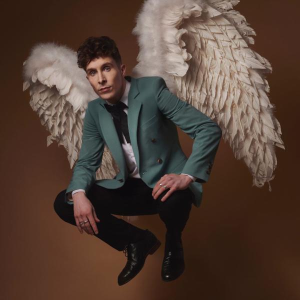 Larry Dean crouches in a suit with a turquoise jacket, resting his hands on his knees and wearing large angel wings on his back.