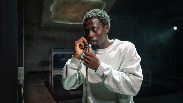 A man in a prison cell is stood holding a phone to his ear.  